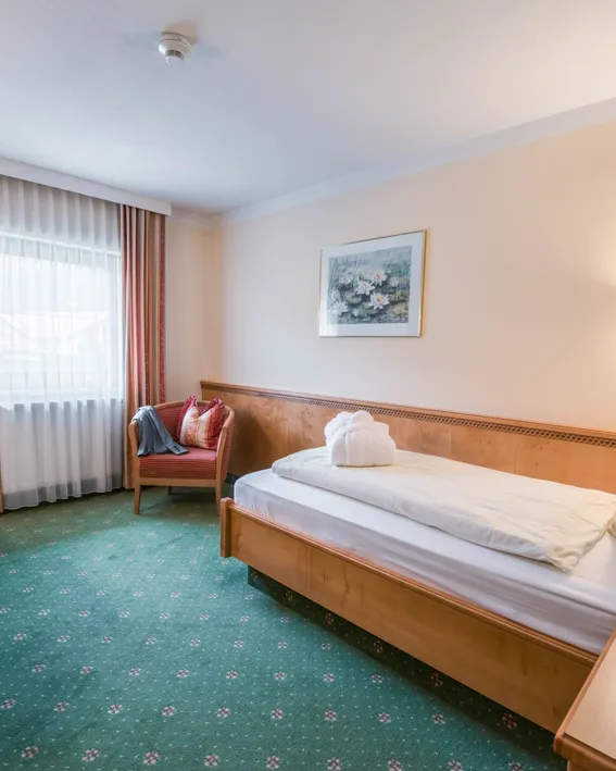 Gemütliches Hotel-Einzelzimmer mit Holzmöbeln, grünem Teppich und Schreibtisch bei Tageslicht
